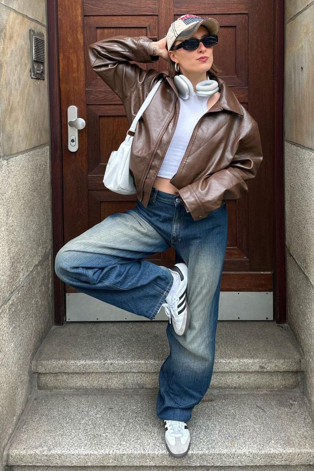 Jaqueta de couro marrom, regata branca, calça baggy jeans, boné estampado e adidas samba
