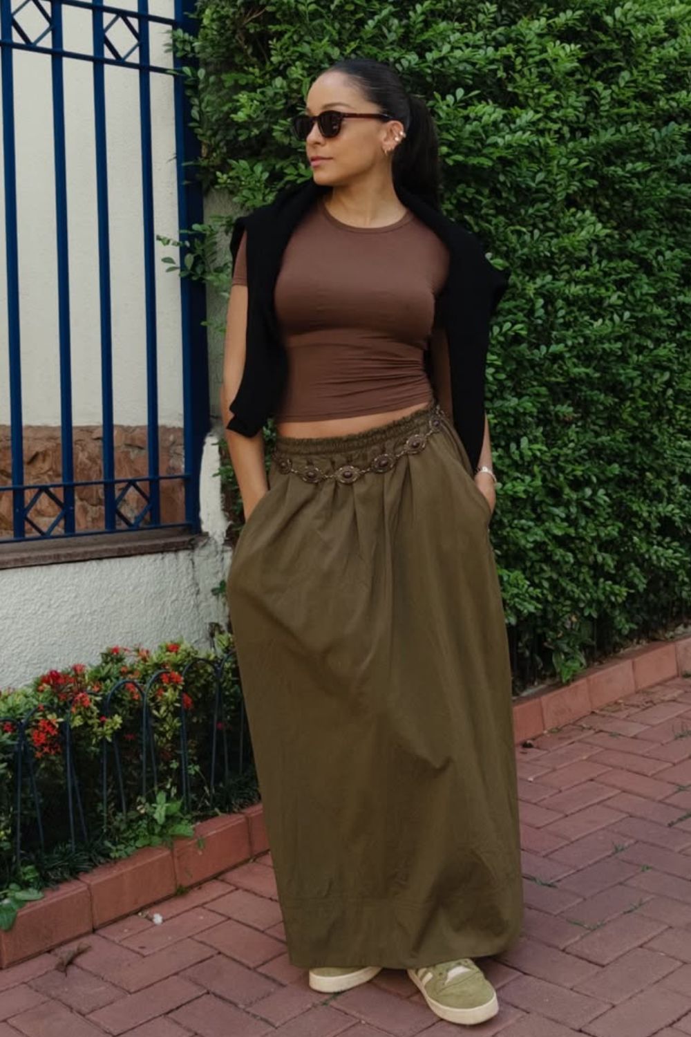 Looks para testar neste outono, blusa marrom, saia longa verde militar e suéter jogado sobre os ombro. Como acessórios: cinto boro e tênis adidas